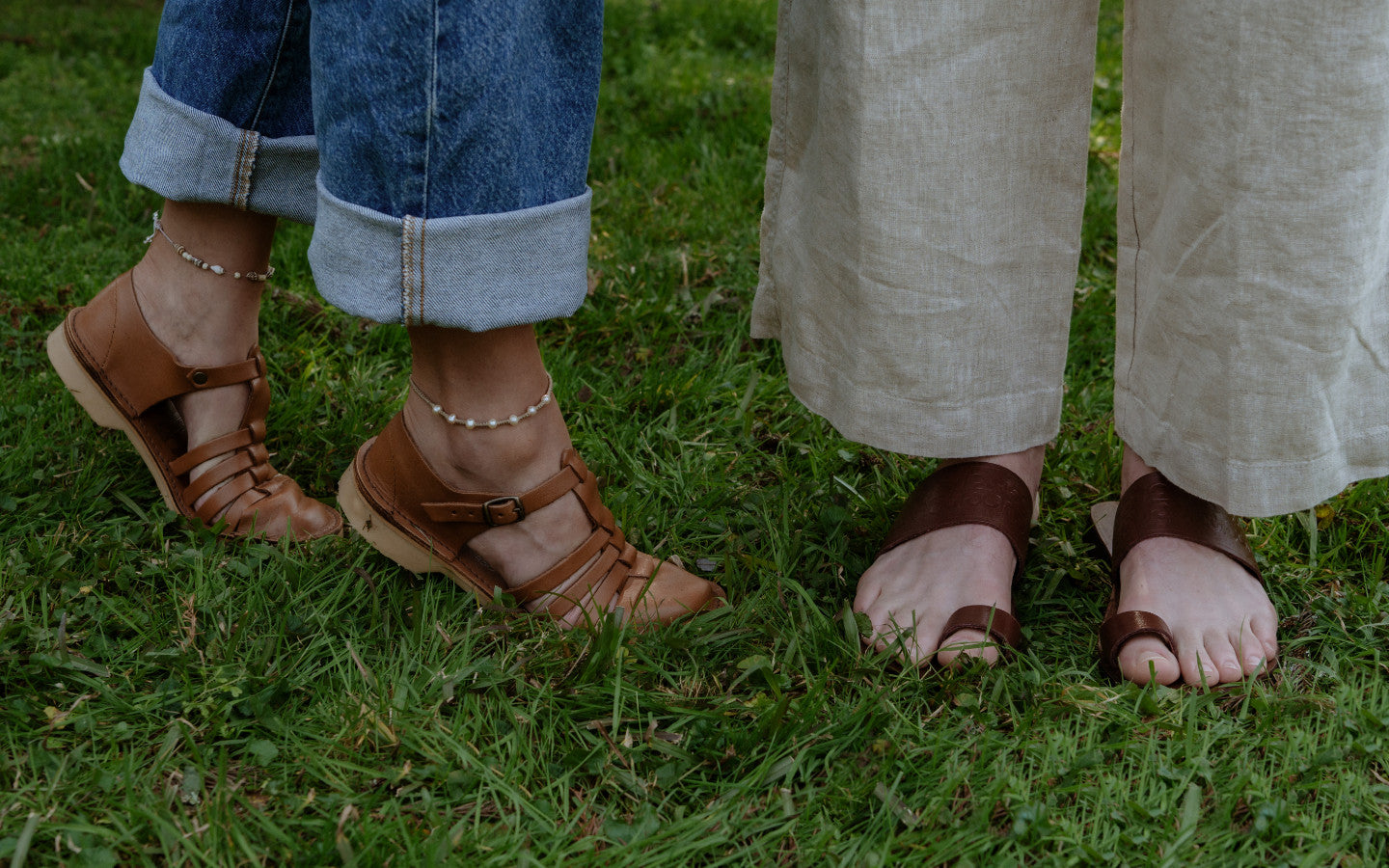 Two pairs of feet wearing brown learher sandals from Groundcover Leather Company on a grassy lawn outdoors.