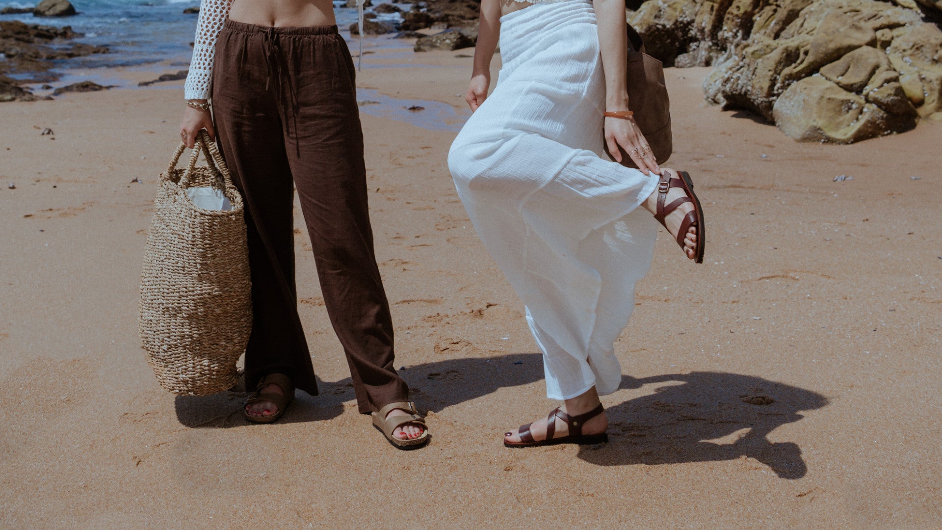 Two people on a beach wearing handmade leather Sandals from Groundcover Leather Comapny with one holding a woven bag.