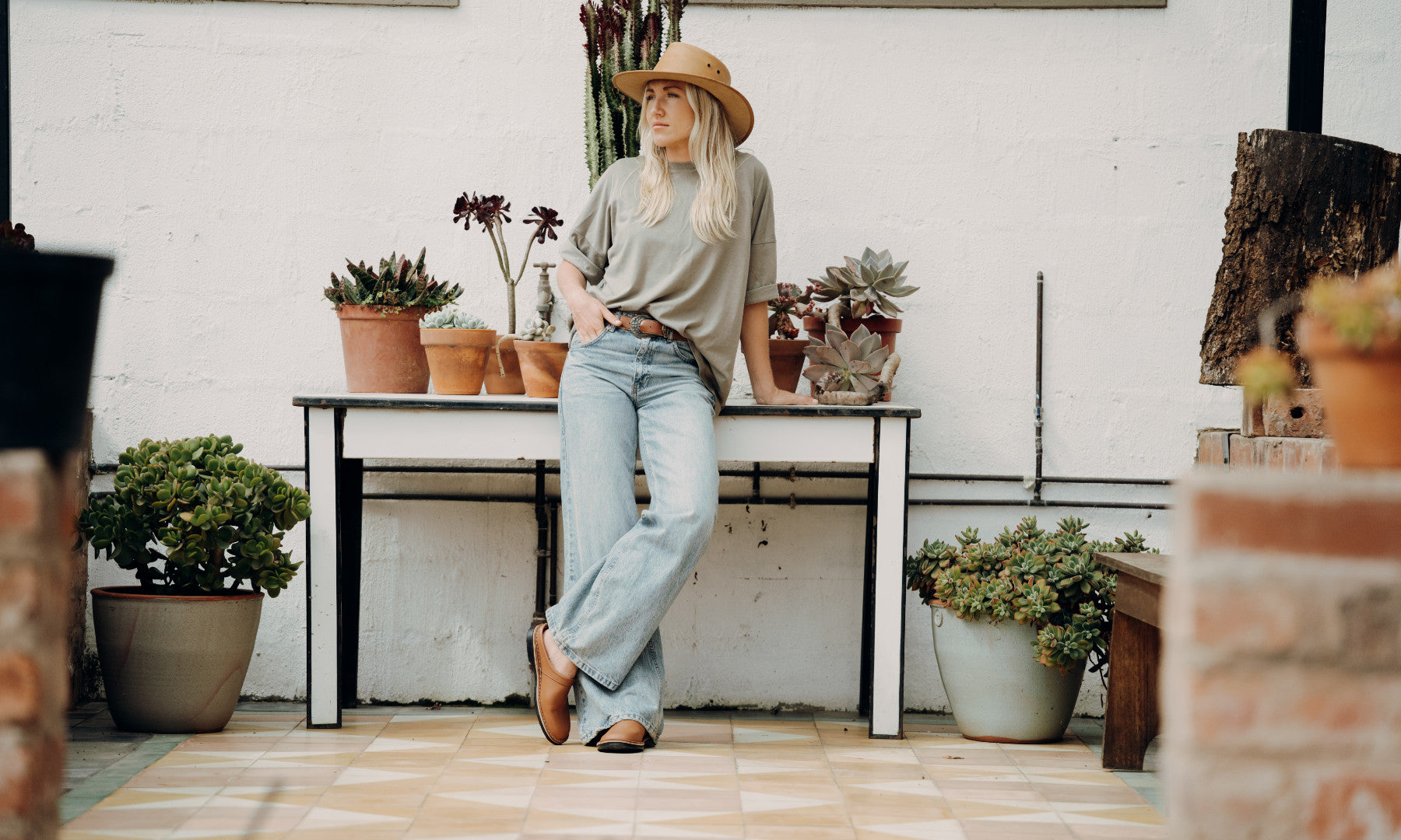 A woman wearing a leather hat, a beige long-sleeve top, light blue wide-leg jeans, and brown handmade leather shoe stands leaning against a white table in a rustic patio setting with potted succulent plants.